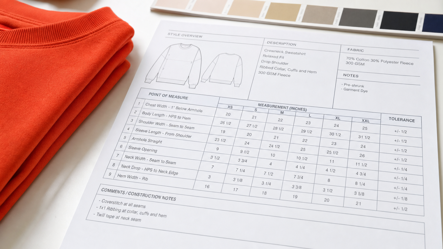 Close-up of a printed graded tech pack measurement table with multiple size columns and tolerance values on a bright desk beside a garment sample and Pantone chips