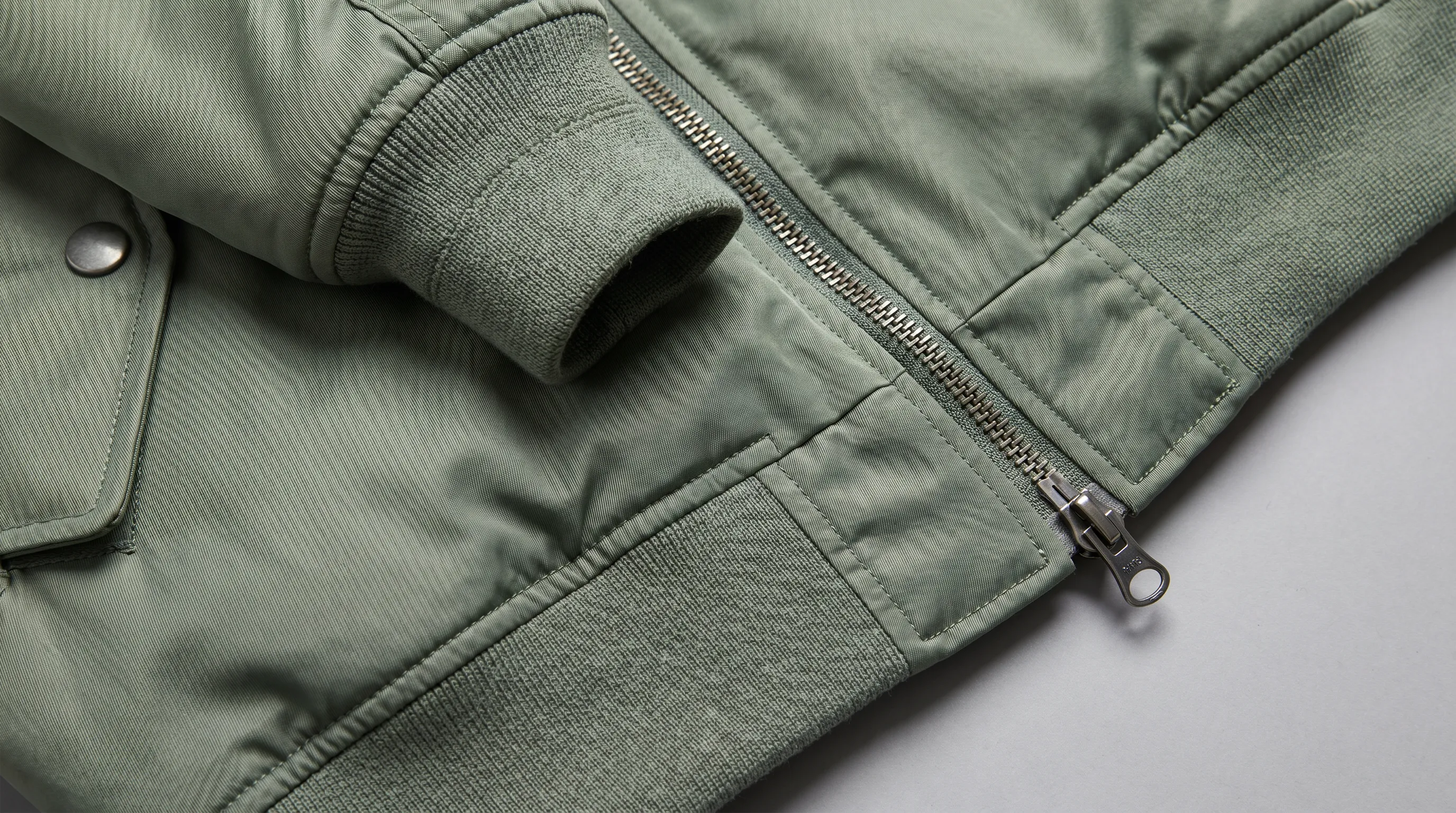 Close-up of bomber jacket tech pack construction details showing the 2x2 ribbed knit cuff, a two-way head-to-head zipper at the hem, and a bartacked snap pocket