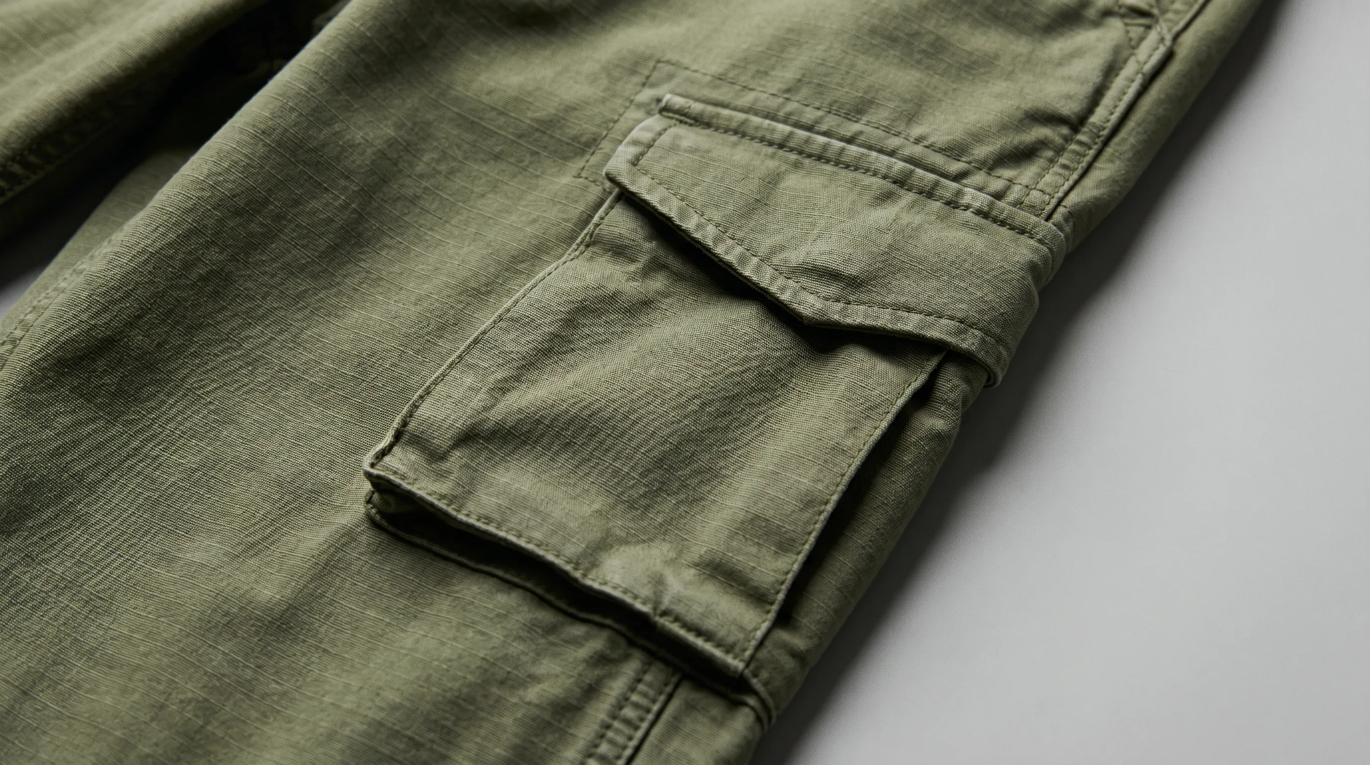 Close-up of cargo pants construction callouts showing a bellows side pocket with a slanted button flap, bartack reinforcements at the corners, and felled outseam topstitching on olive ripstop