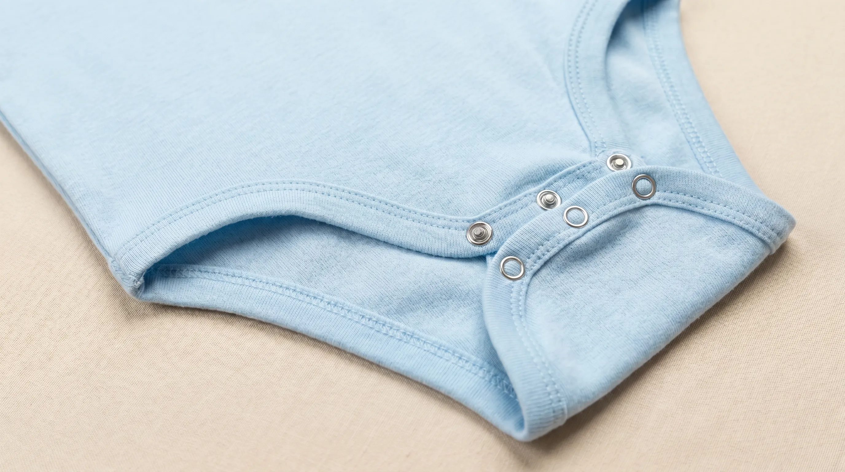 Macro close-up of a toddler snap-front bodysuit showing reinforced shoulder snaps, a snap-crotch inseam, and flat covered seams on a clean neutral surface